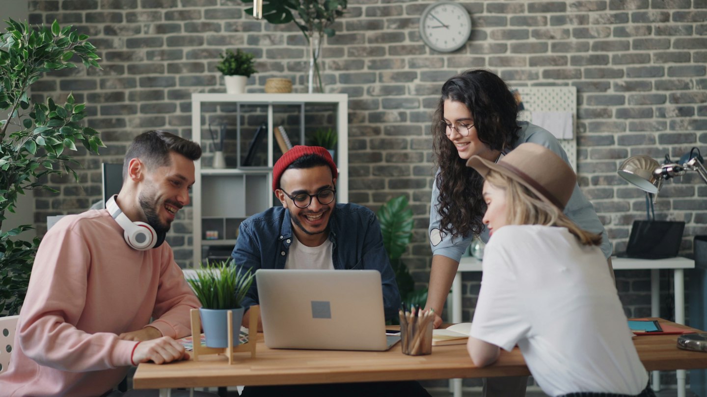 Four young professionals smiling and collaborating around a laptop at a desk.