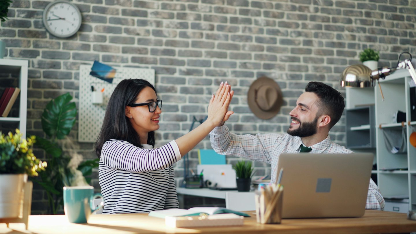 Two colleagues high-fiving at a wooden desk in a modern office.