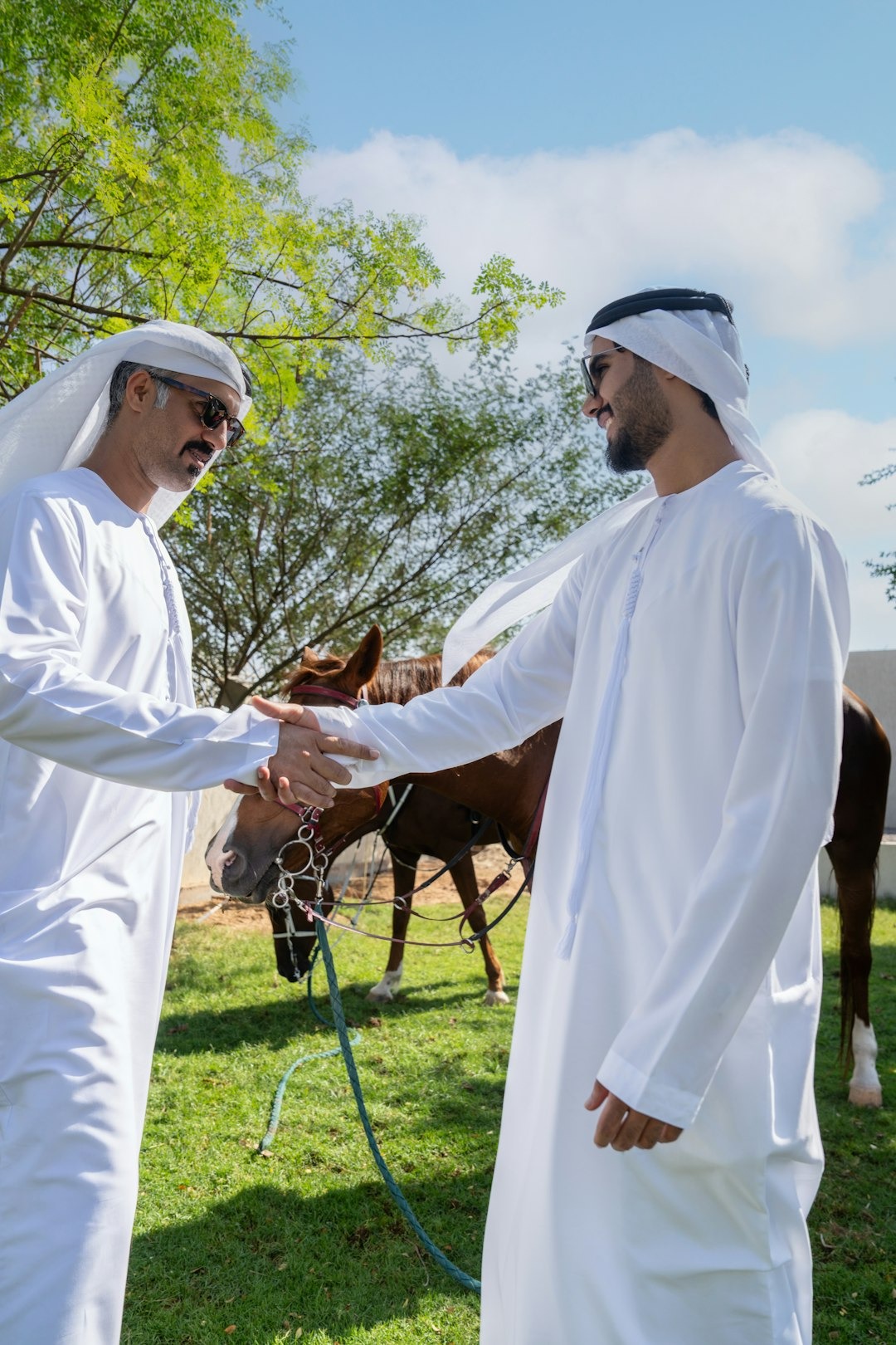 Two men in white thobes shake hands near a saddled horse.